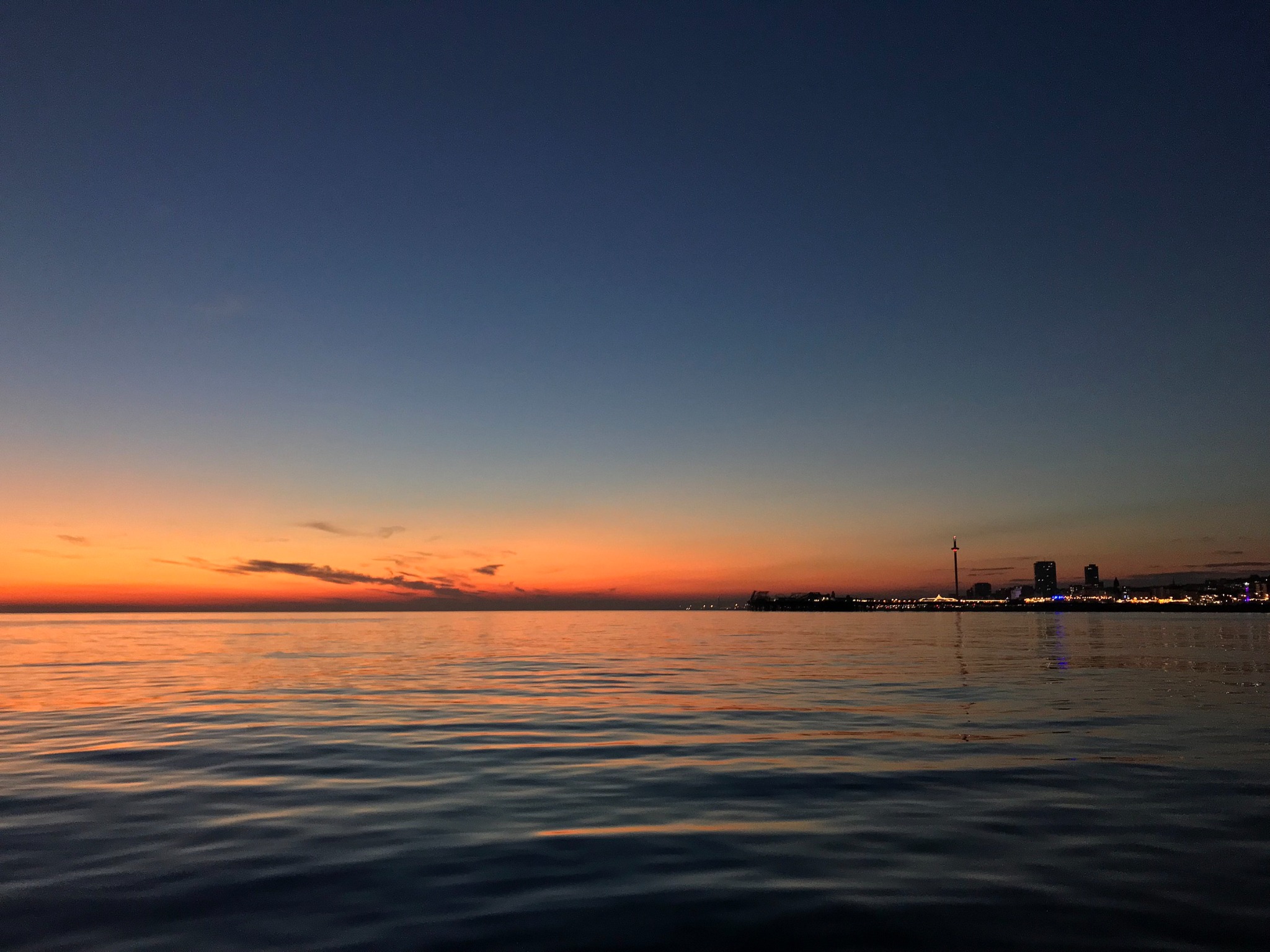 Brighton seafront at sunset, with calm water, glowing horizon light, and the distant city skyline.